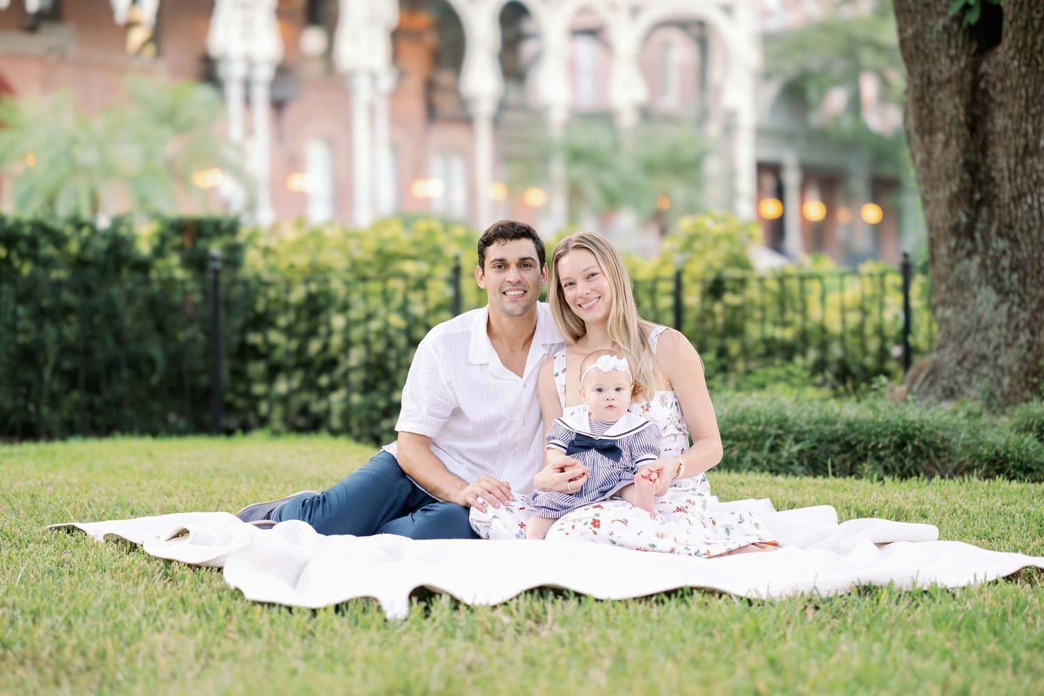 Young family with baby sitting on white blanket on grass in park, smiling at camera on sunny day.