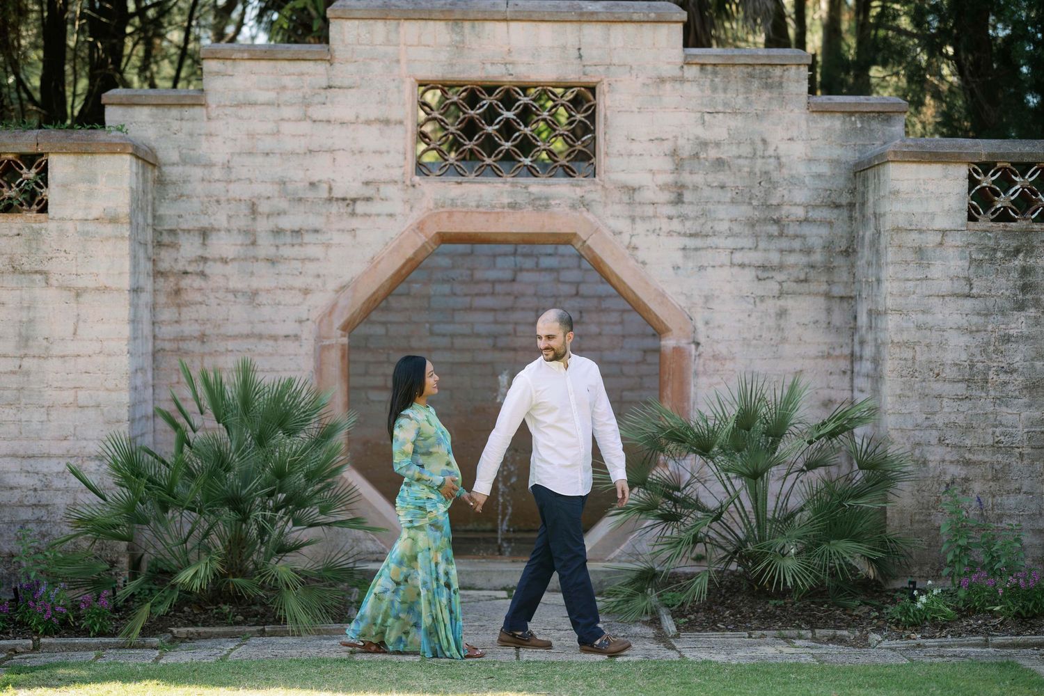 Couple holding hands walking in front of an ornate stone garden wall with circular window and tropical plants.