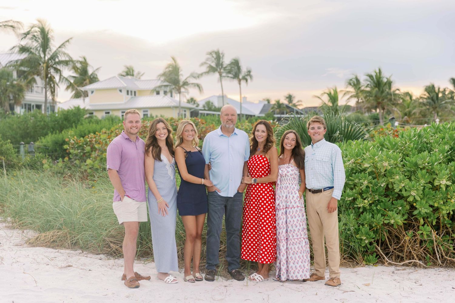 Family of seven posing together on a sandy beach at sunset with palm trees and tropical greenery in the background.