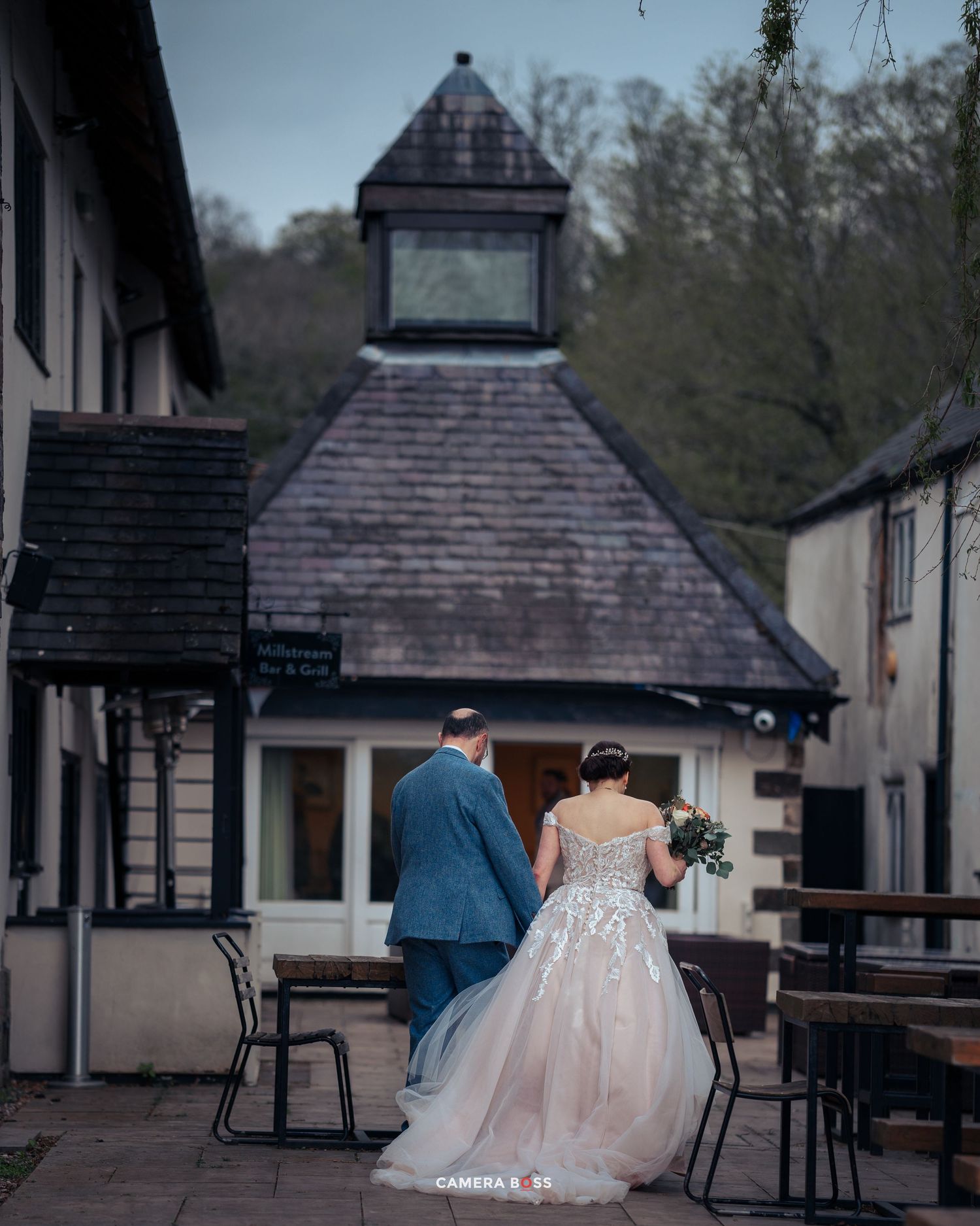 A couple in wedding attire stand together on a patio with outdoor seating and a rustic gazebo-style structure in the background.