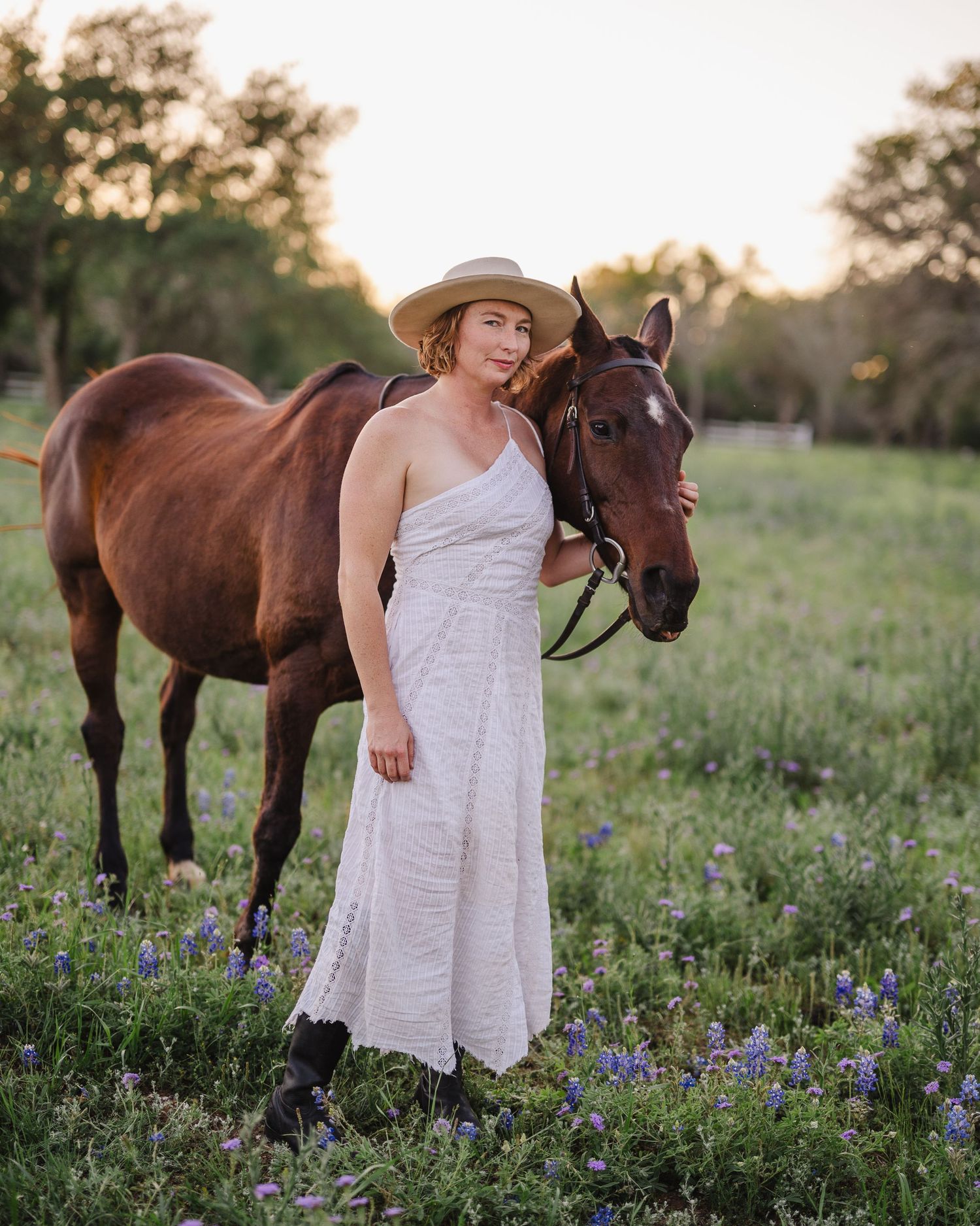 Woman in white dress poses with brown horse in field of spring wildflowers.