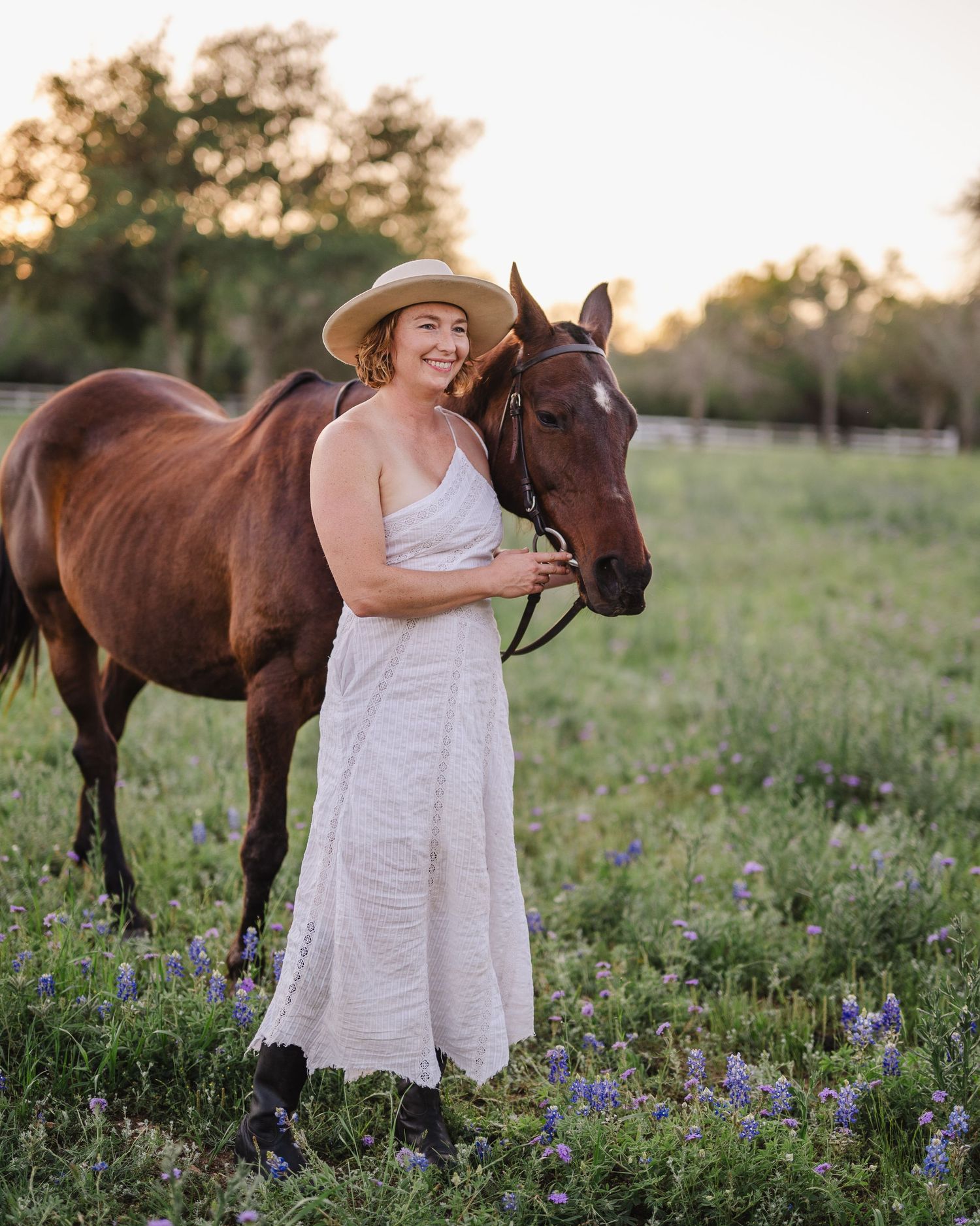 A person in a white dress and sunhat stands lovingly with their horse in a field of bluebonnets at sunset.