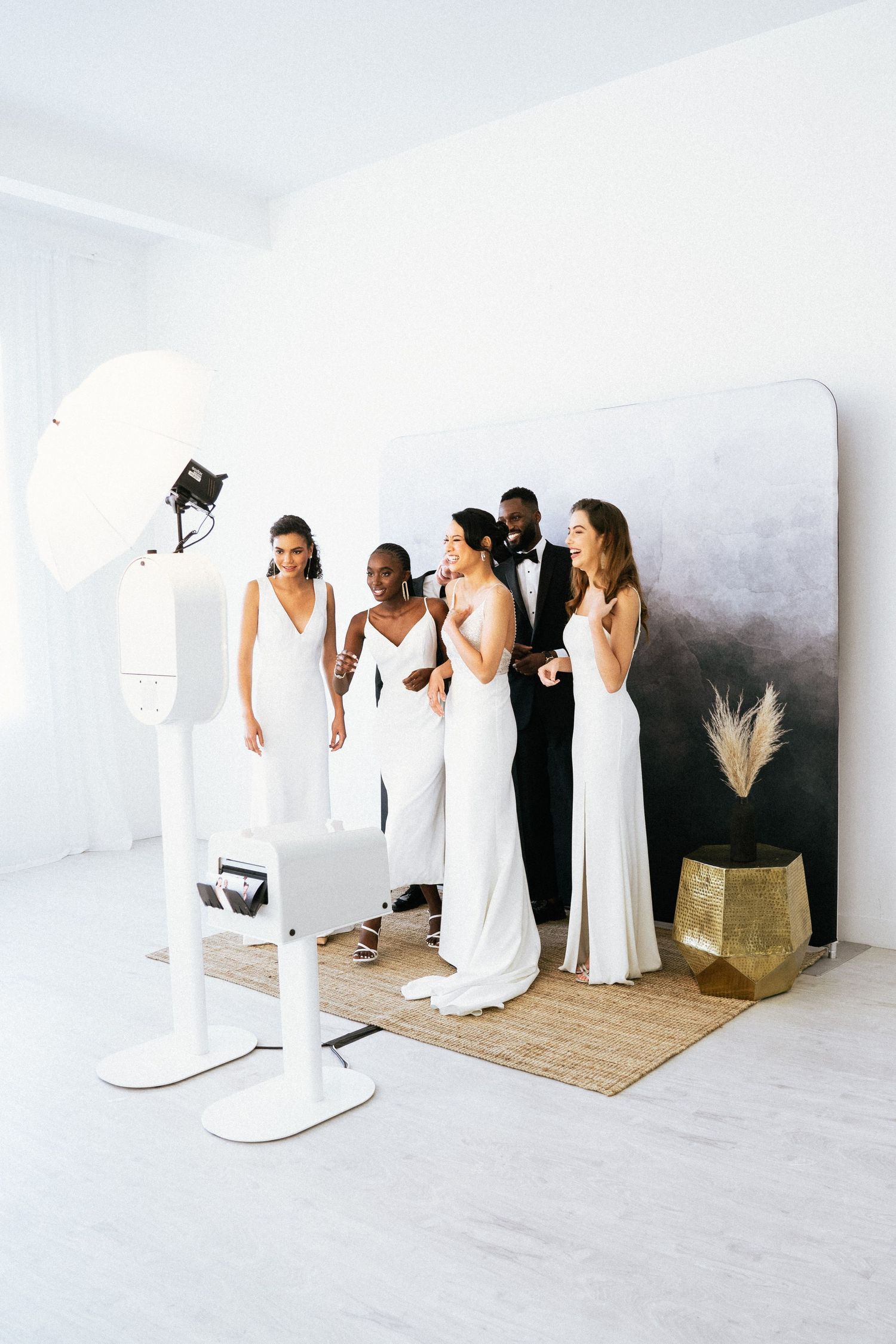 A group in white and black dresses poses against a white wall in a minimalist studio setting using a photobooth.