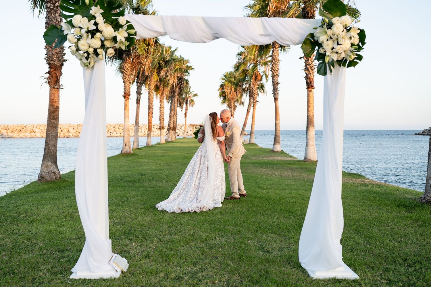 An elegant beach wedding ceremony setup with white draped arch and palm trees overlooking the ocean.