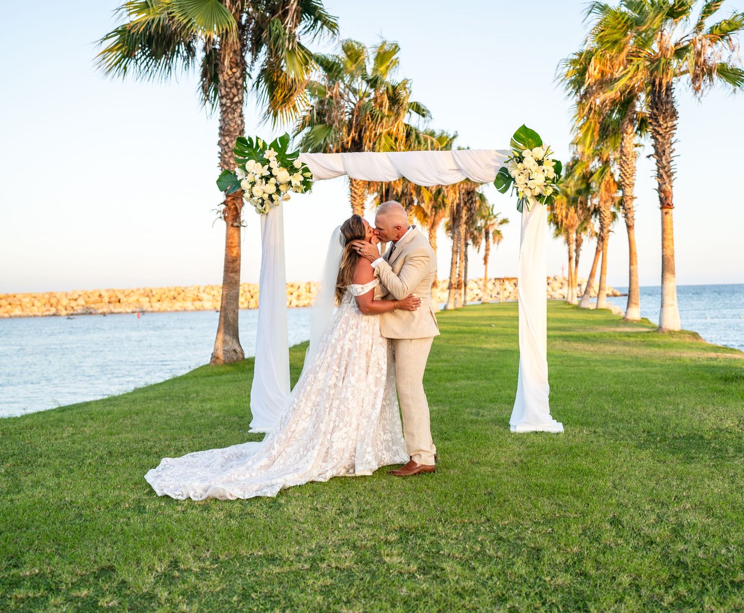 A newlywed couple shares a moment under a decorated arch with palm trees and ocean views.