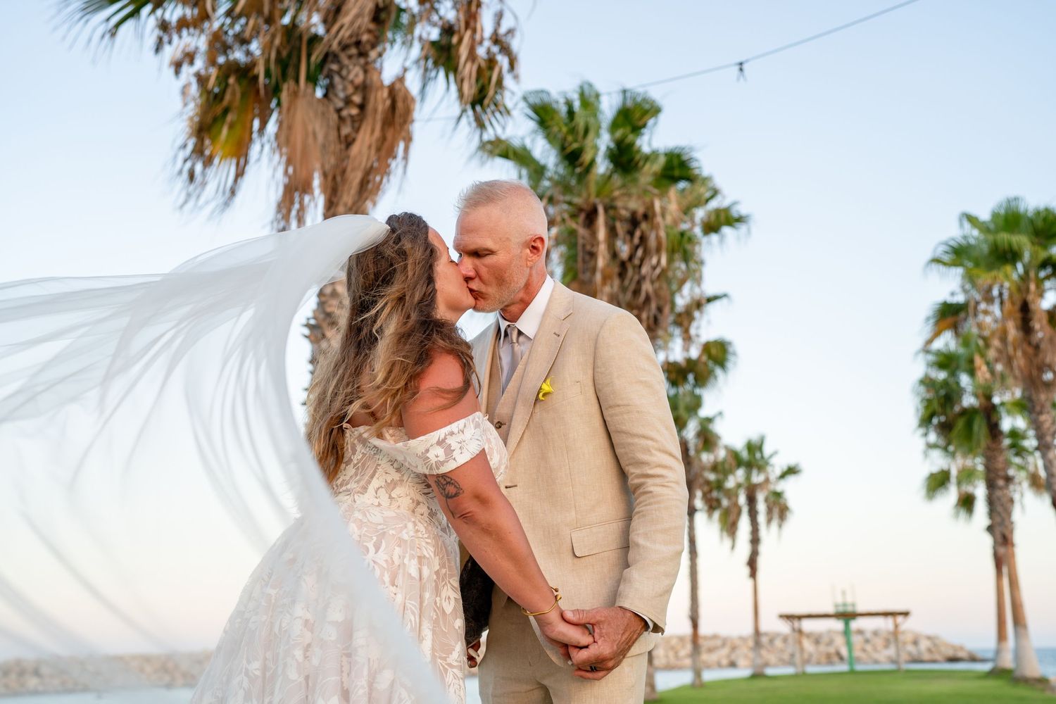A romantic wedding photo with palm trees along a beach as a flowing veil catches the breeze.