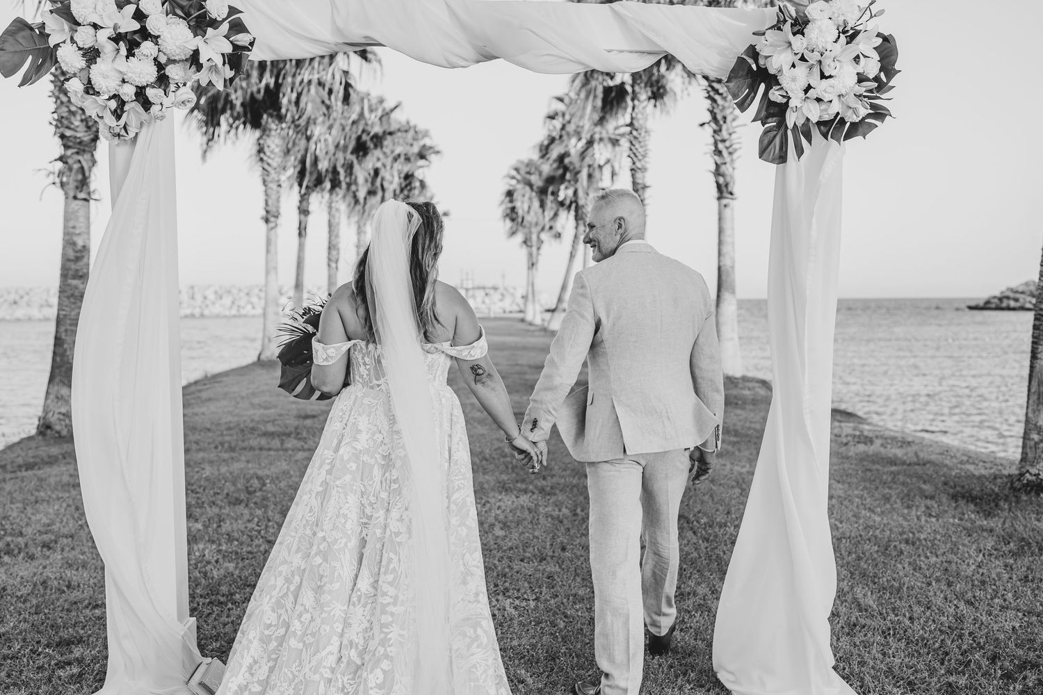 A wedding ceremony under a floral arch with white draping by the ocean.