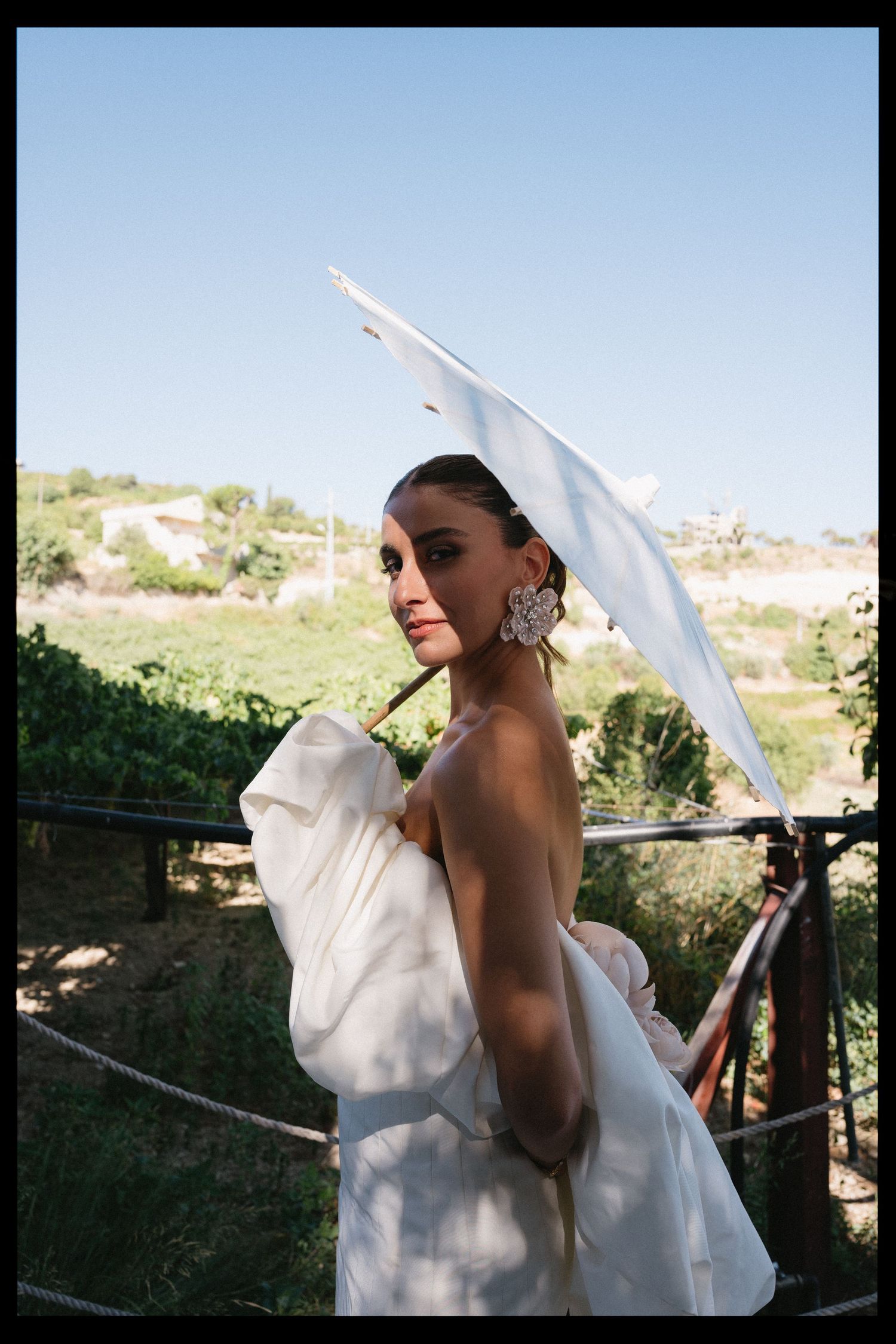 A person in an elegant white dress poses with a parasol against a scenic hillside backdrop.