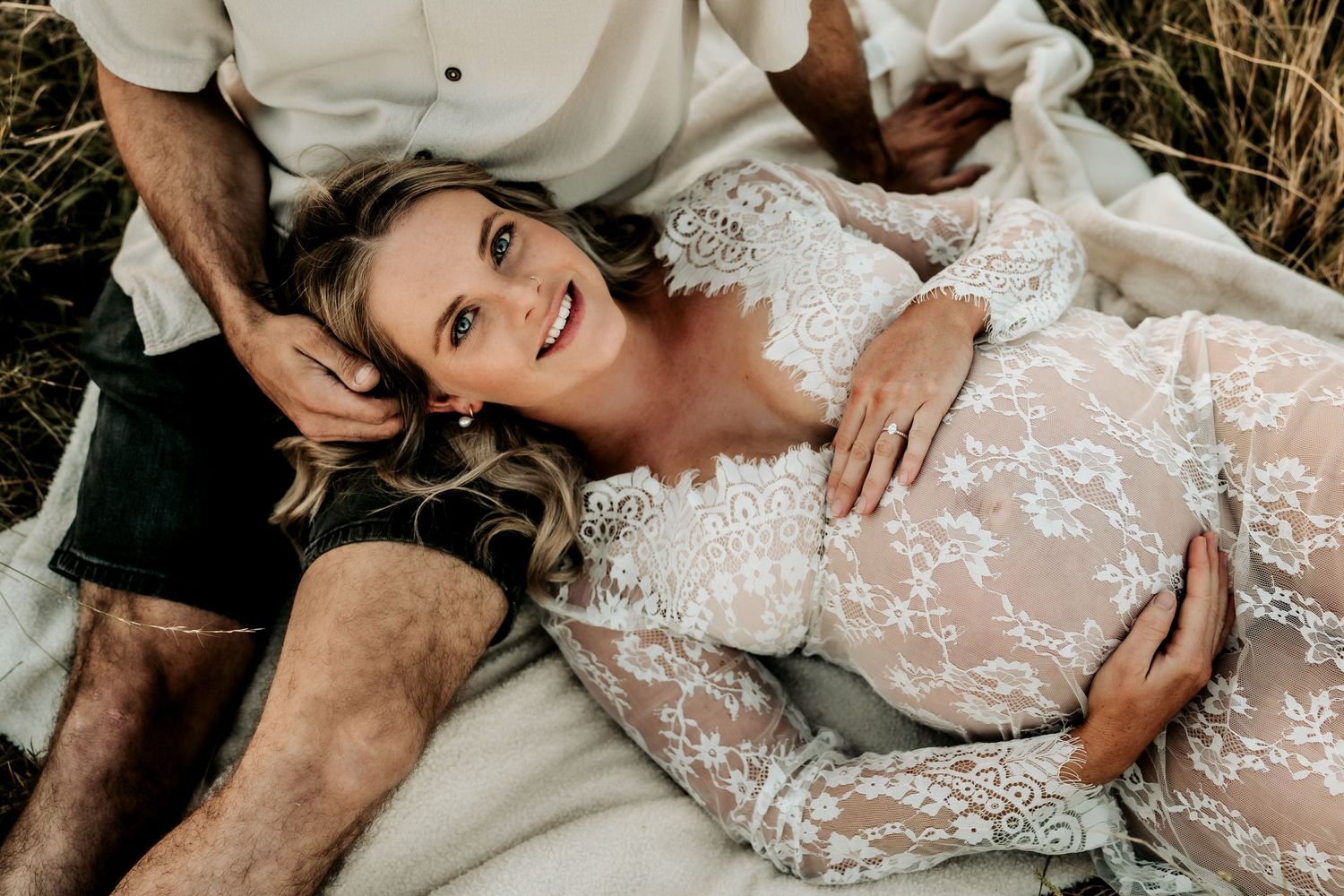 Pregnant woman in lace gown lying in grass with partner during golden hour maternity shoot.