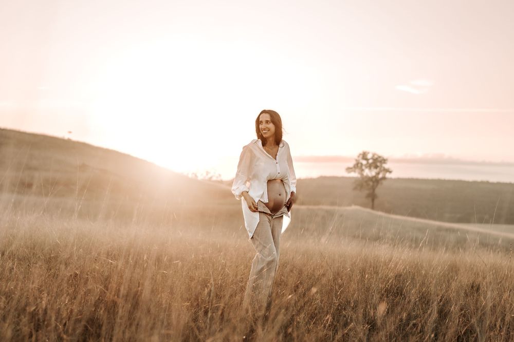 Pregnant woman in white outfit walking through sun-drenched golden field at sunset.