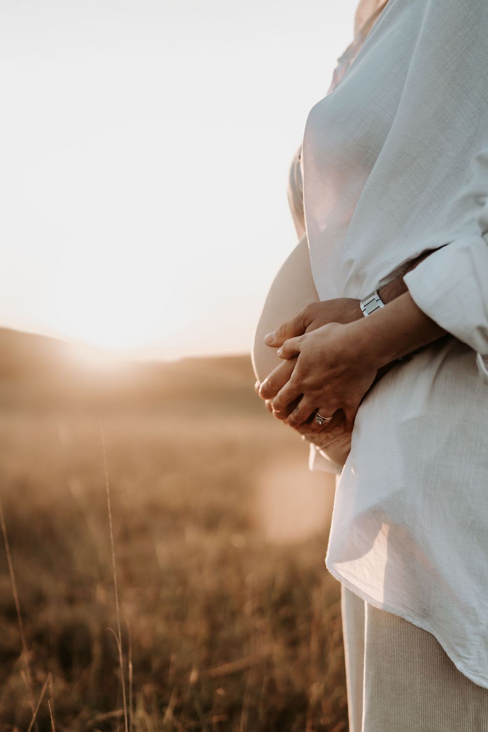 Close-up of pregnant woman cradling baby bump in white shirt with partner's hands at golden sunset.