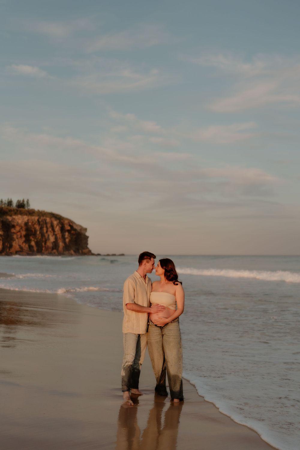 Pregnant couple embracing on wet sandy beach at sunset with rocky coastal cliffs in background.