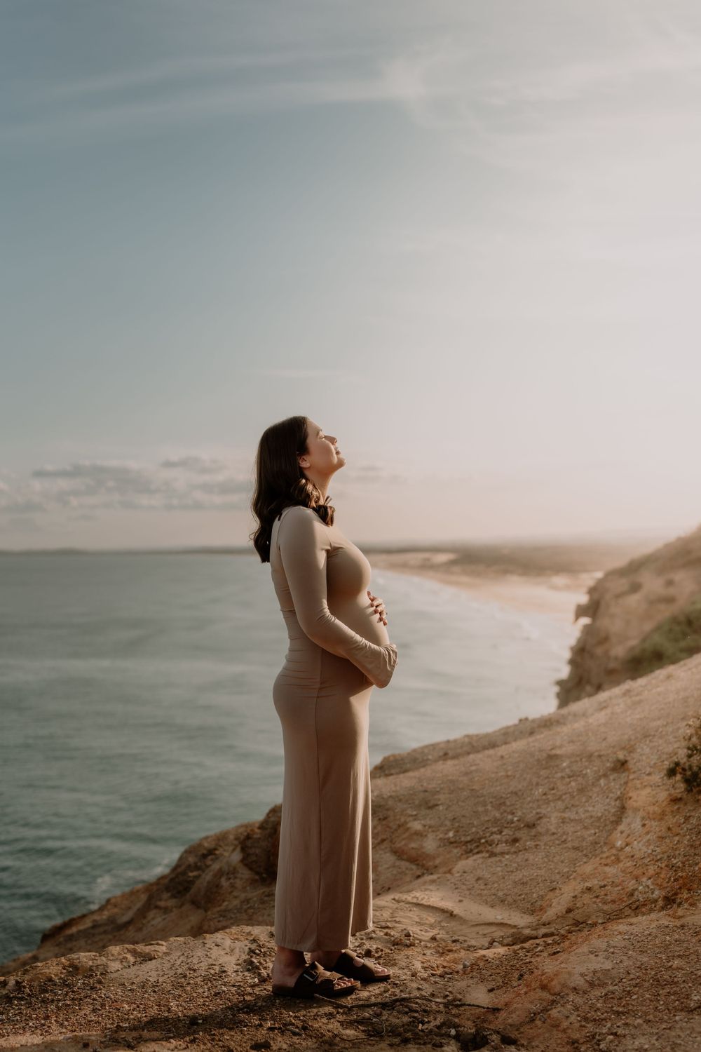 Pregnant woman in beige dress standing on coastal cliff, cradling bump, ocean view behind her.