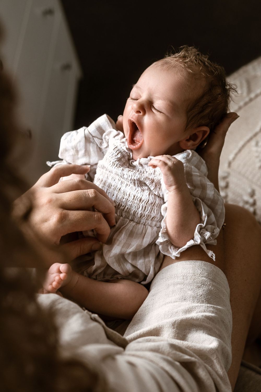 Newborn baby yawning while being held by curly haired mother in warm light.