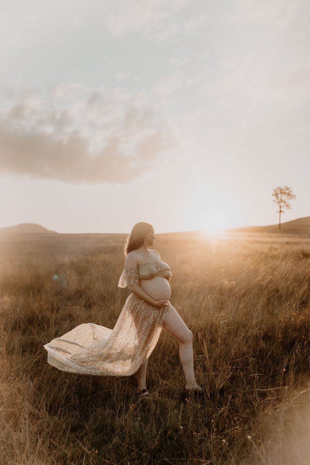 Pregnant woman in flowy lace dress walking through golden grass field at glowing sunset.
