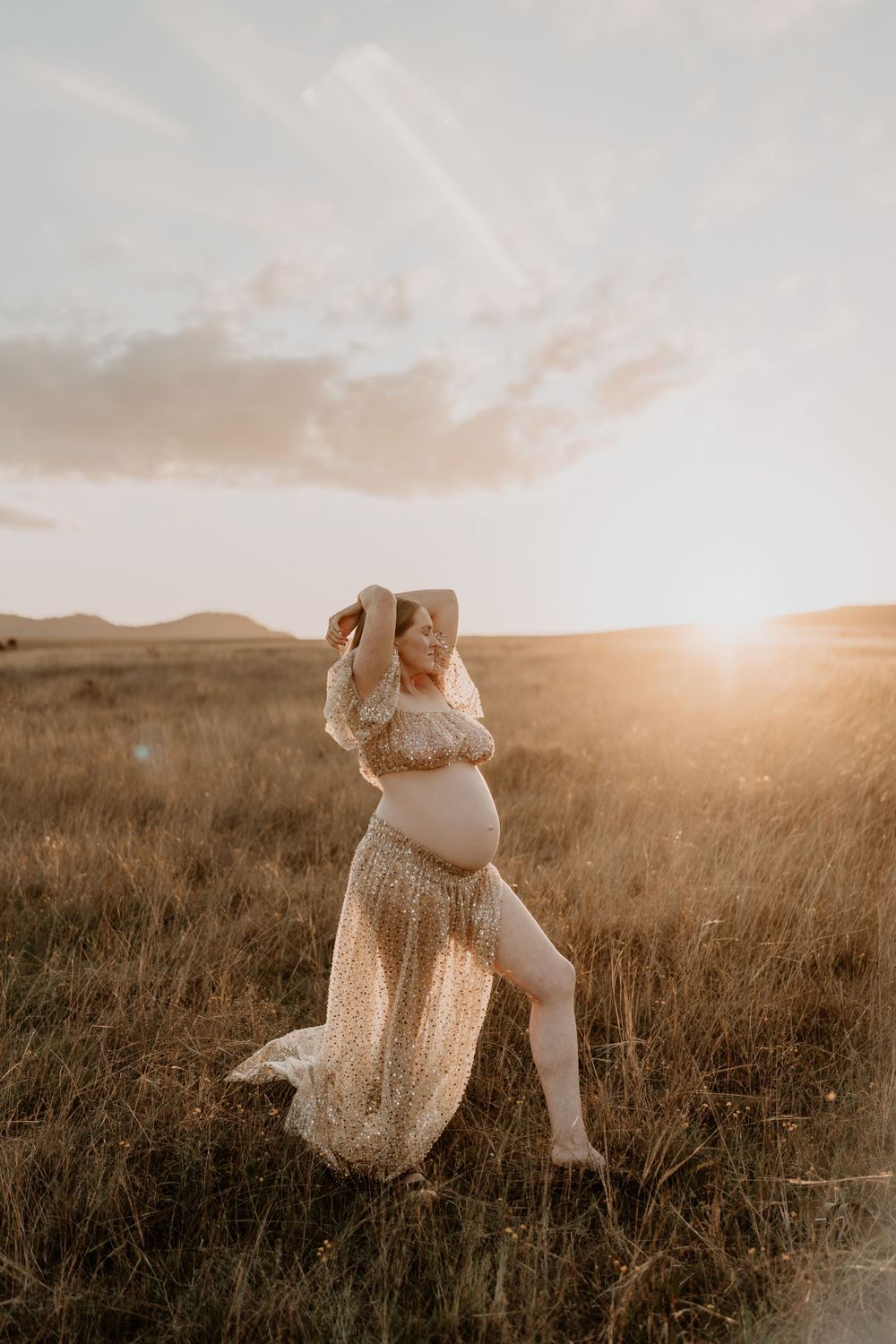 Expecting mother in sheer lace maternity gown posing barefoot in sunlit golden meadow at sunset.