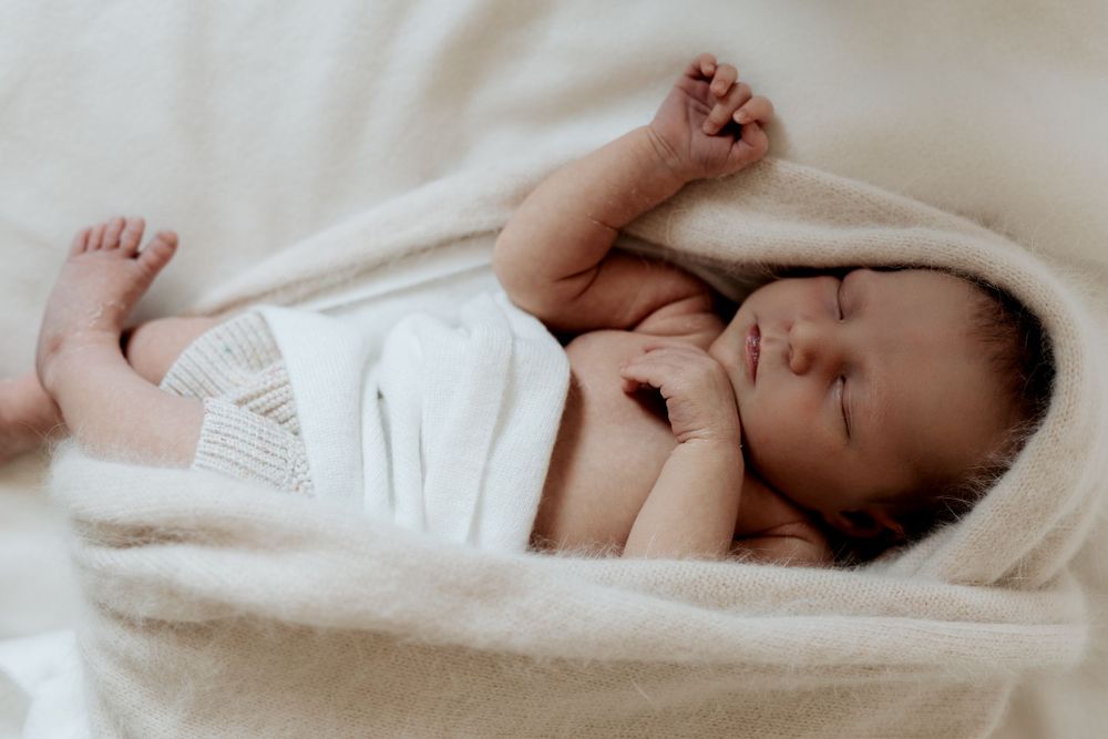 Peaceful sleeping newborn baby wrapped in soft white blanket with tiny fist raised.