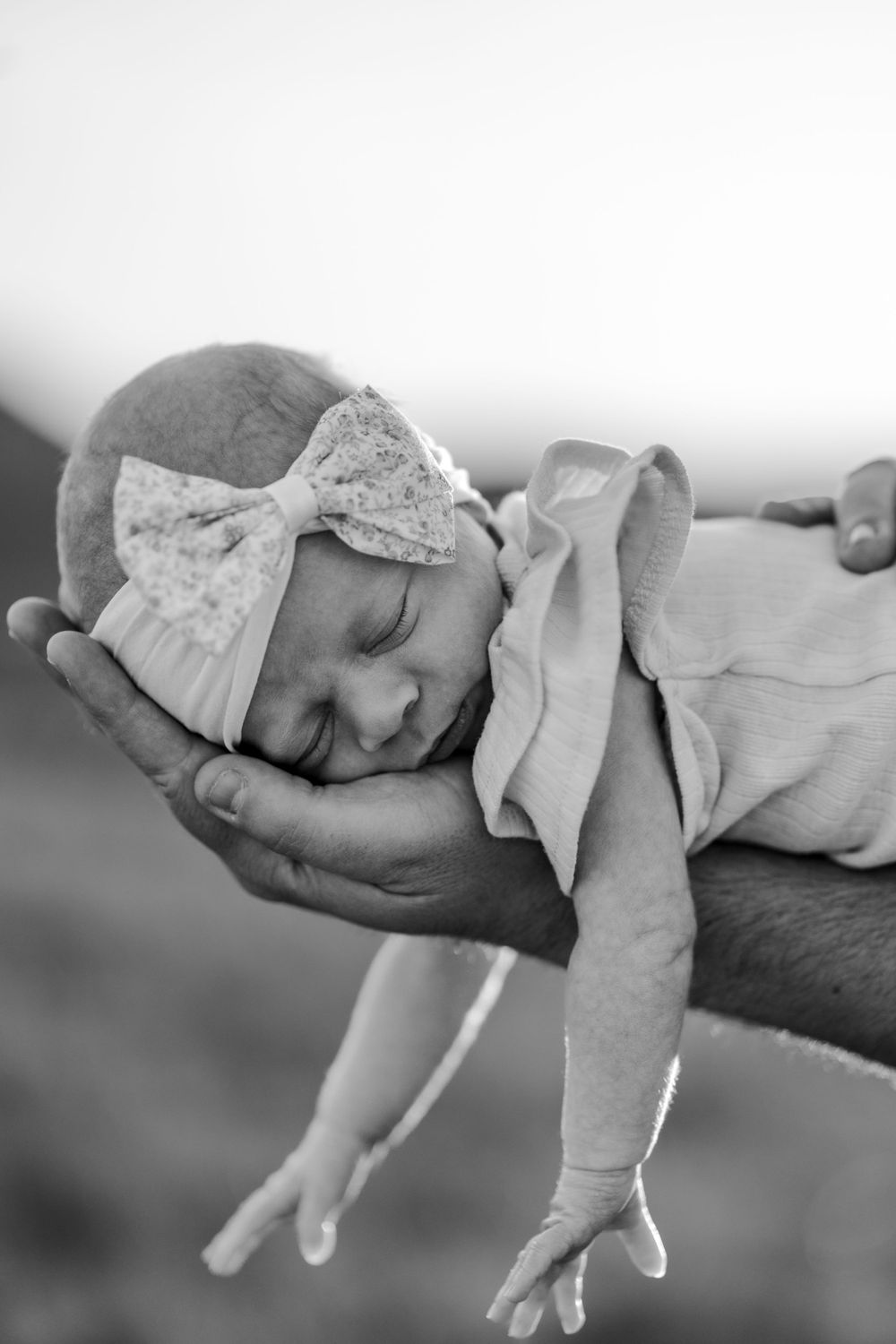 Black and white photo of sleeping newborn baby girl with bow headband cradled in hands.