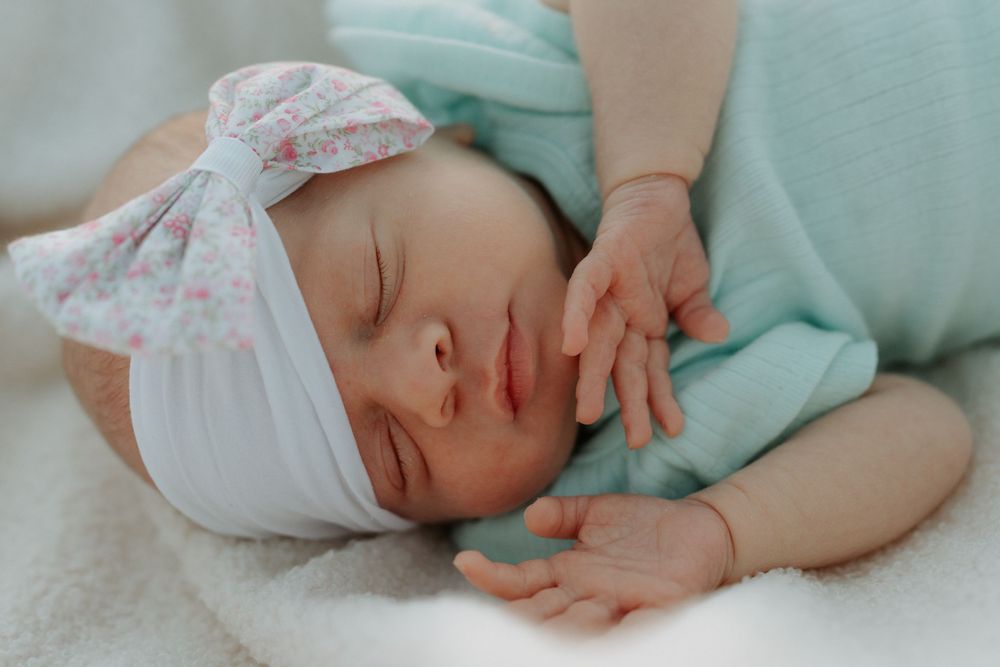 Peaceful sleeping newborn baby girl in mint wrap with floral bow headband on white blanket.