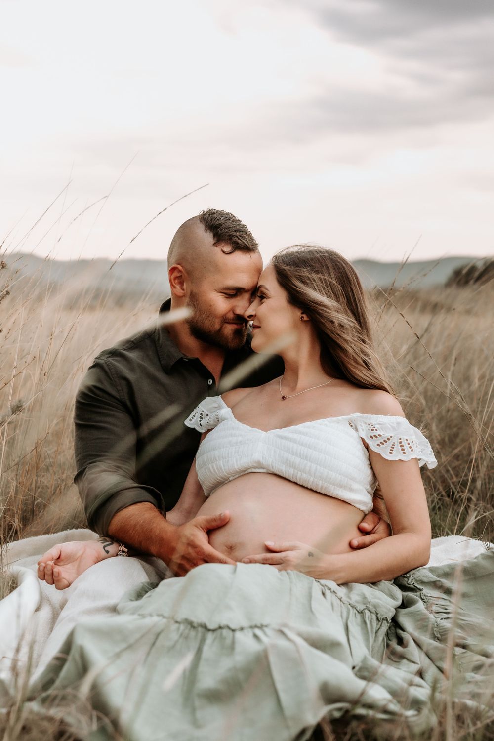 Couple sitting in golden field, man kissing pregnant woman as she cradles her baby bump.
