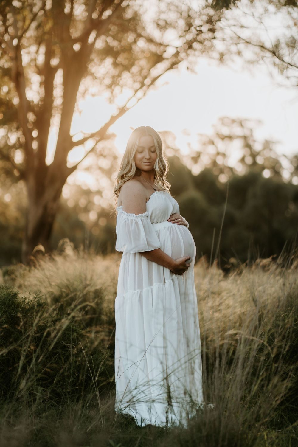 Pregnant woman in white off-shoulder dress cradling baby bump in golden sunlit field.