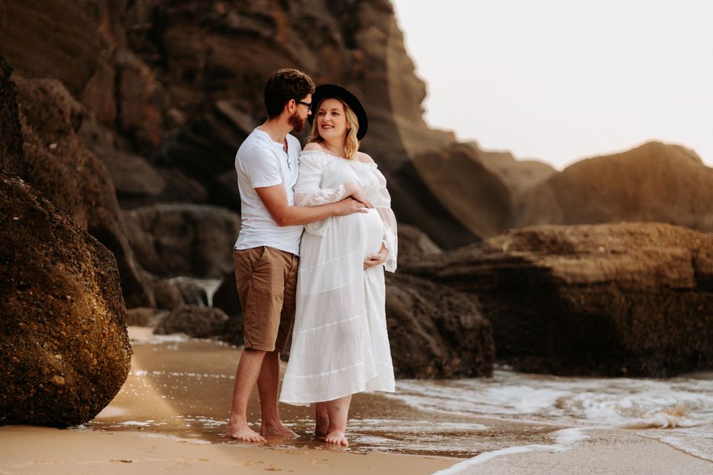 Couple embracing on a sandy beach with rocky backdrop during golden hour maternity photoshoot.