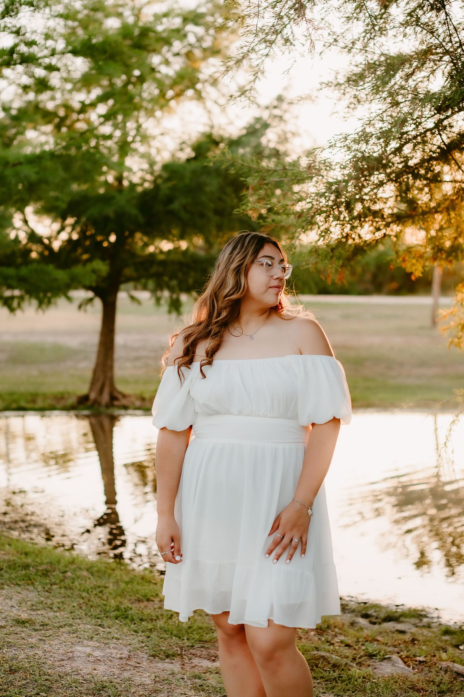 A serene portrait by a pond at sunset wearing a white off-shoulder dress.