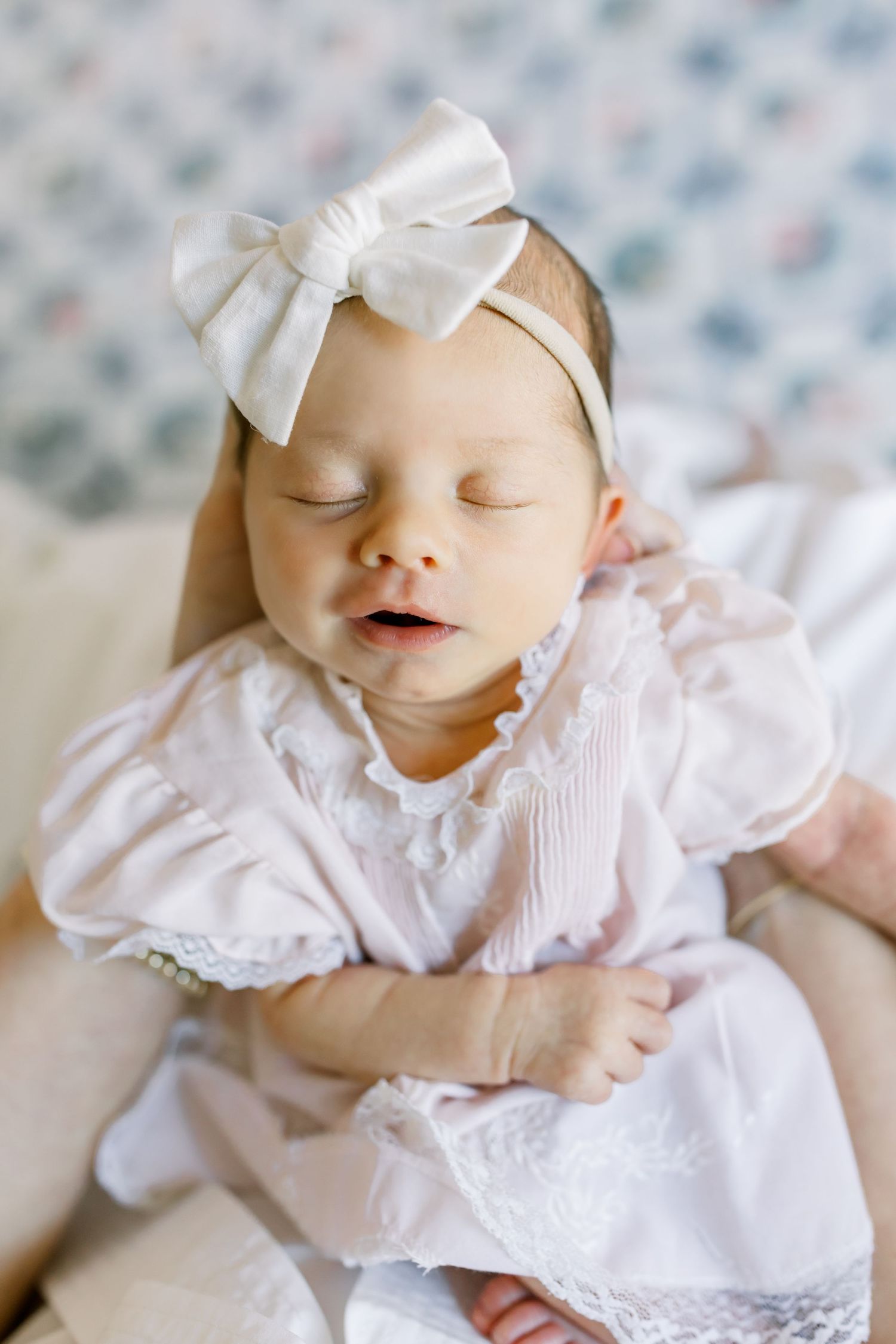 Sleeping newborn wearing white dress and large bow headband against soft blue background.