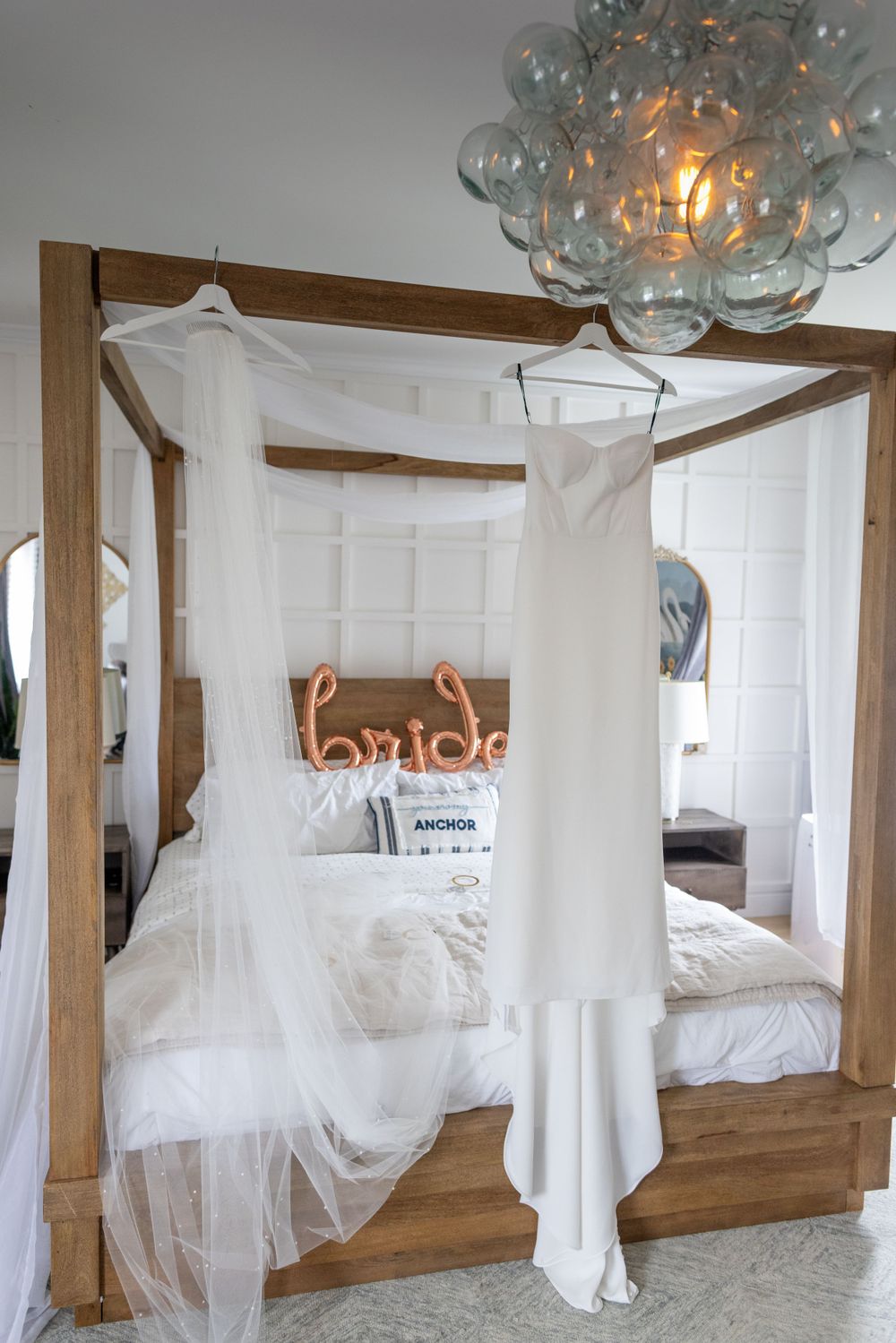 Elegant white wedding gown hangs on wooden four-poster canopy bed with neon bride sign and chandelier.