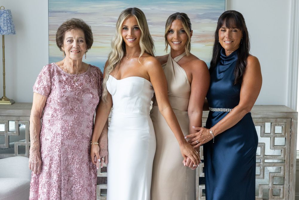 Bride in white gown poses with three women in formal dresses including grandmother in pink lace before wedding.