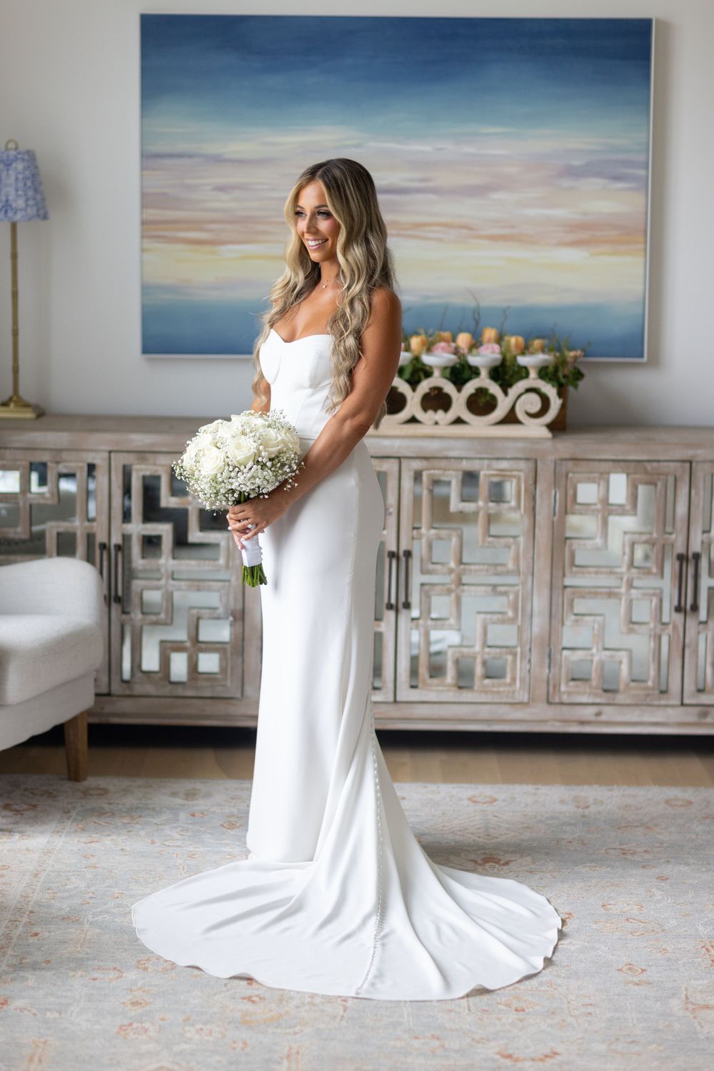 Elegant bride in strapless white gown holds white bouquet standing before colorful abstract painting in bright room.