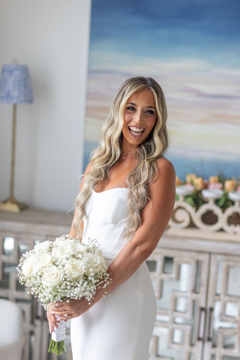 Smiling bride in strapless white dress holds white rose and baby's breath bouquet with long wavy blonde hair.