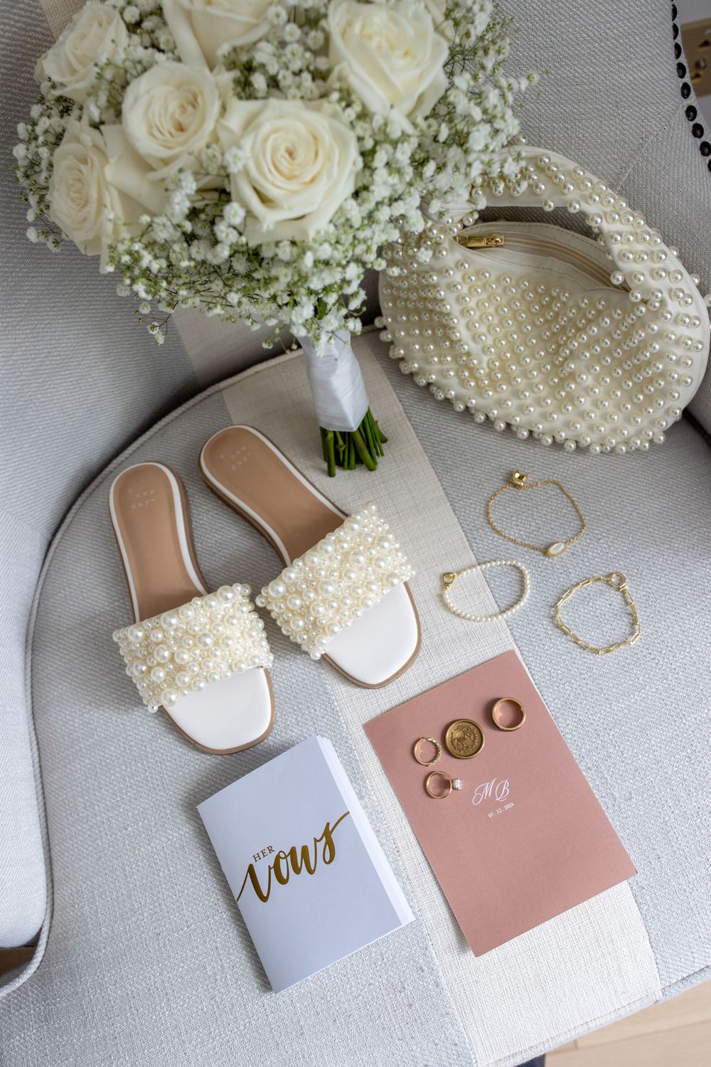 Flat lay of bridal accessories including pearl shoes, bouquet, vow books, rings, and jewelry.