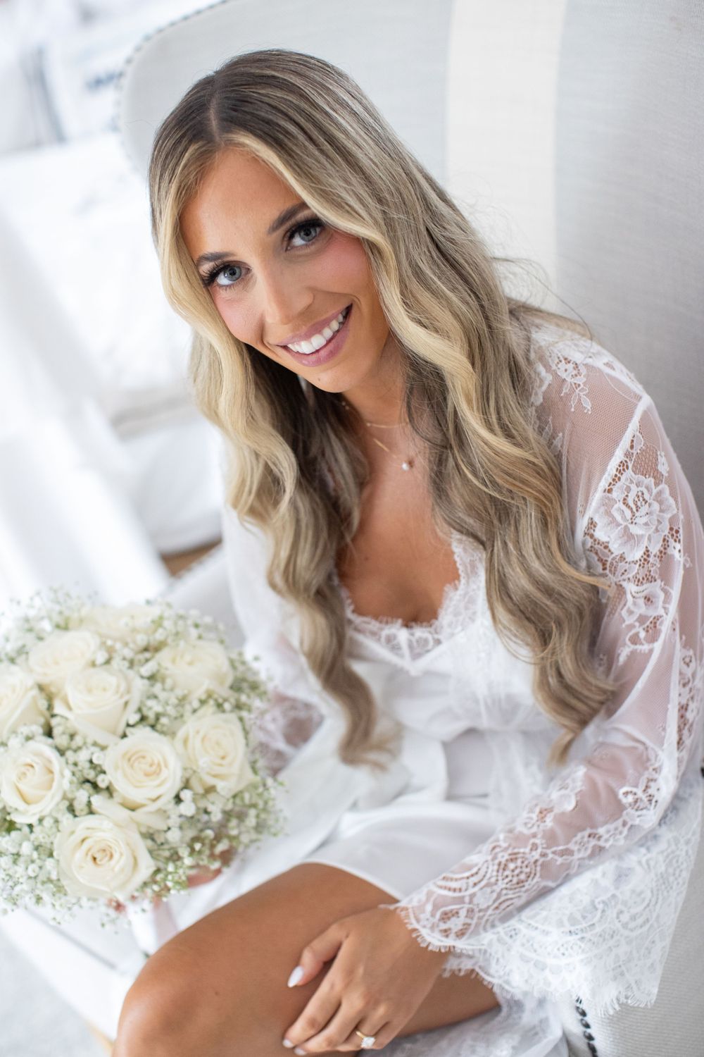 Smiling bride in white lace robe holds a bouquet of white roses before her wedding.