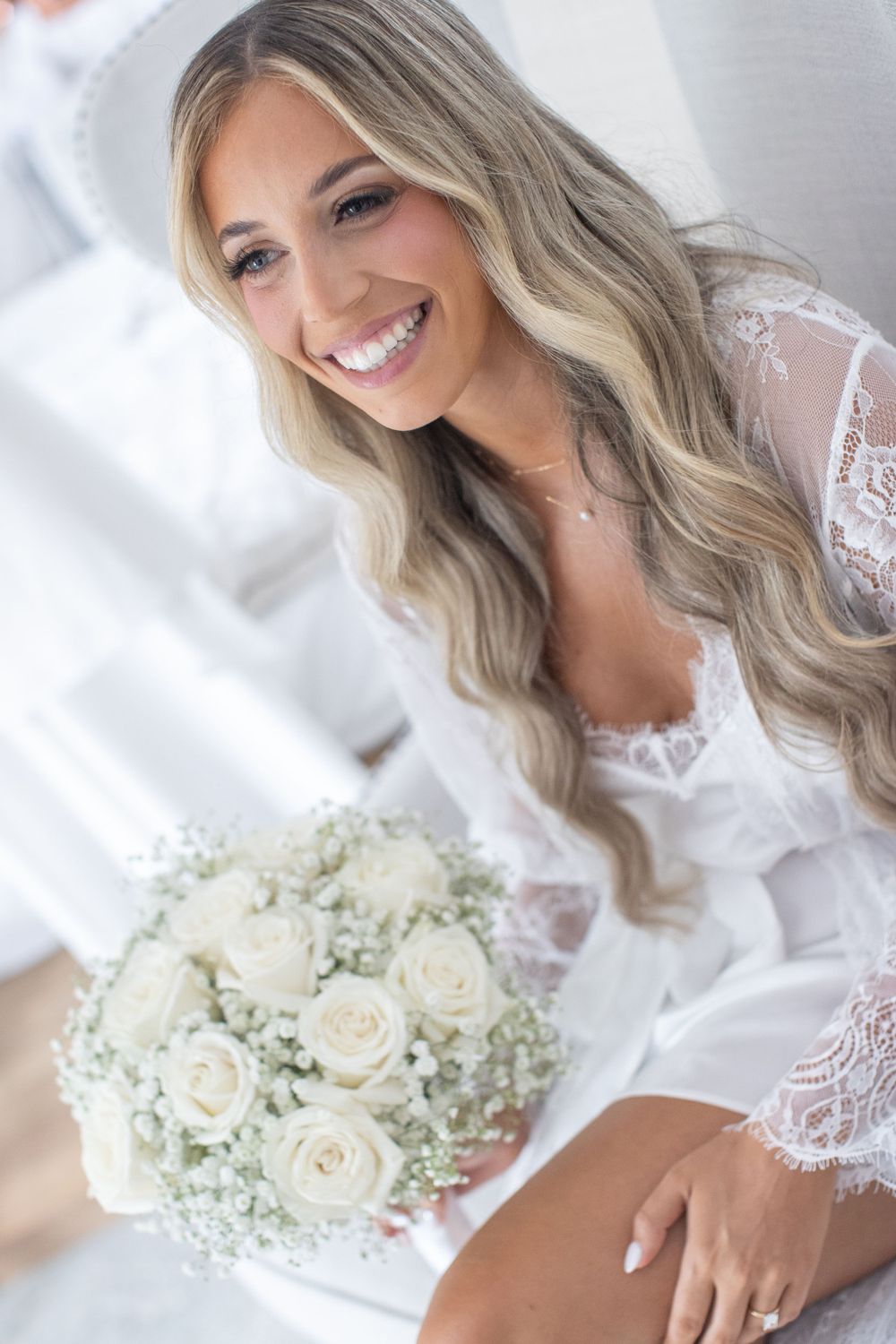 Radiant bride with long blonde waves holds white rose bouquet in a bright bridal suite.