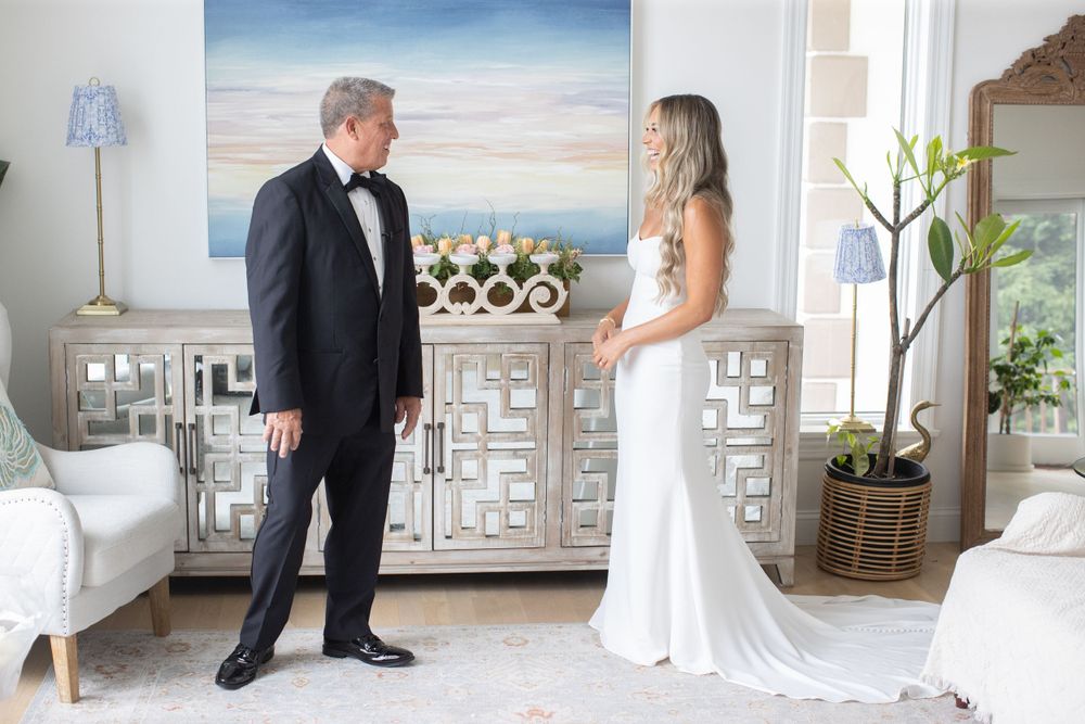 Father in tuxedo shares emotional first look moment with bride in white gown in elegant coastal home.