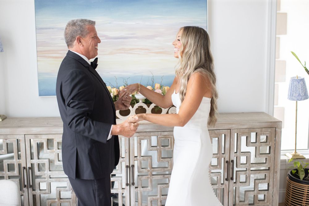 Father and bride hold hands during heartfelt first look reveal in beautifully decorated coastal interior.