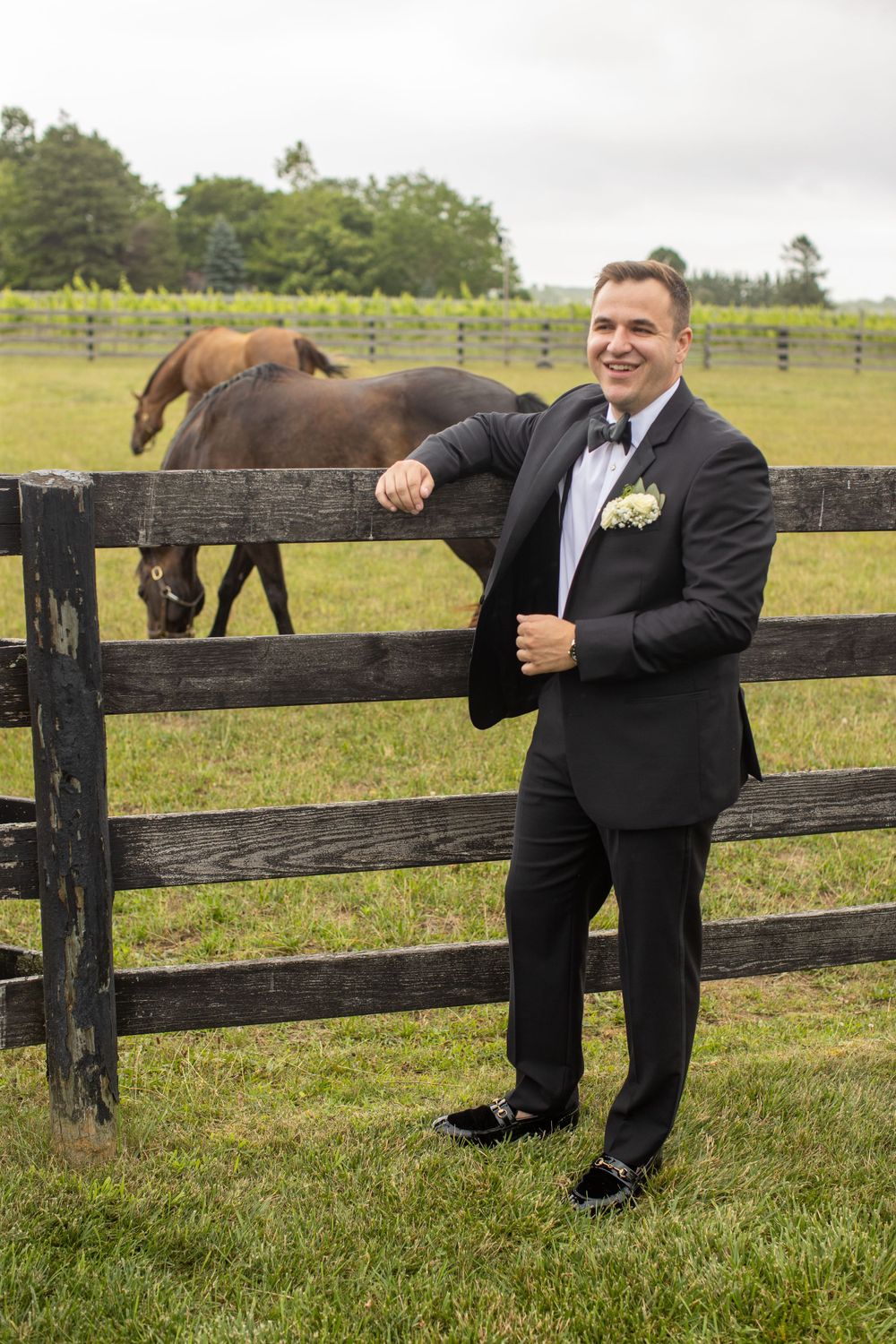 Dapper groom in black tuxedo smiles while leaning on rustic wooden fence with horses in background.