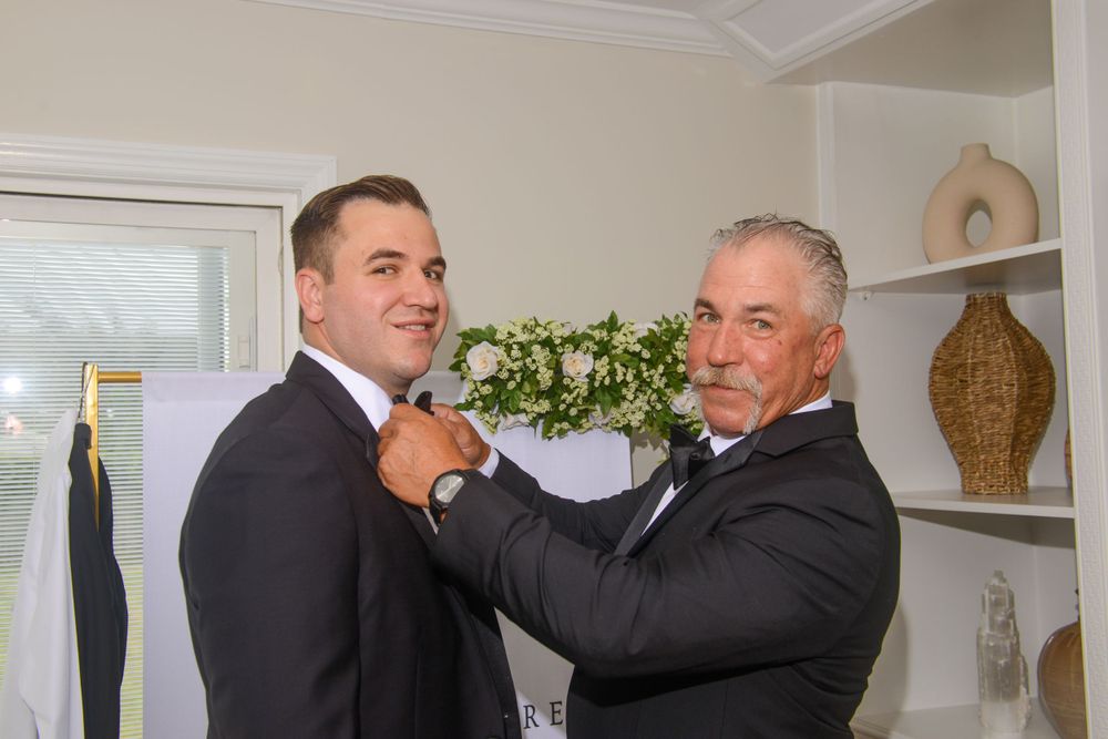 Older man helps groom adjust boutonniere on black tuxedo jacket during wedding day preparations indoors.