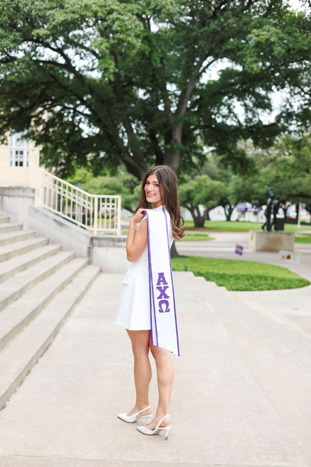 Graduate in white dress celebrates with purple and white tassel on outdoor steps.