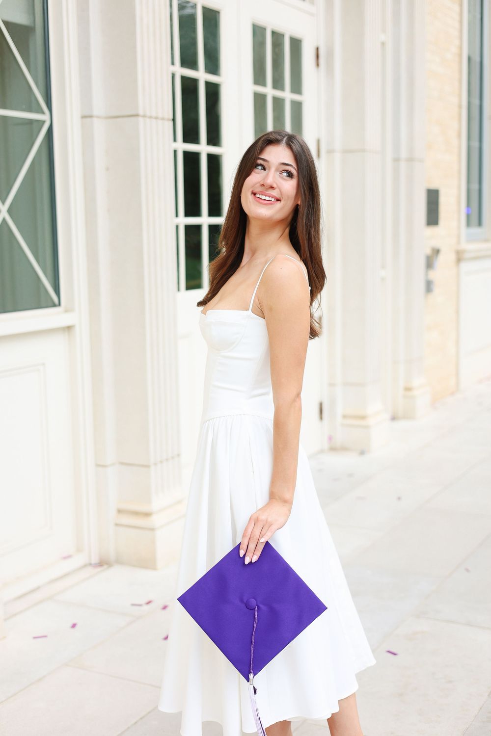 Graduate in white dress holds purple diploma cover while posing by white columns.