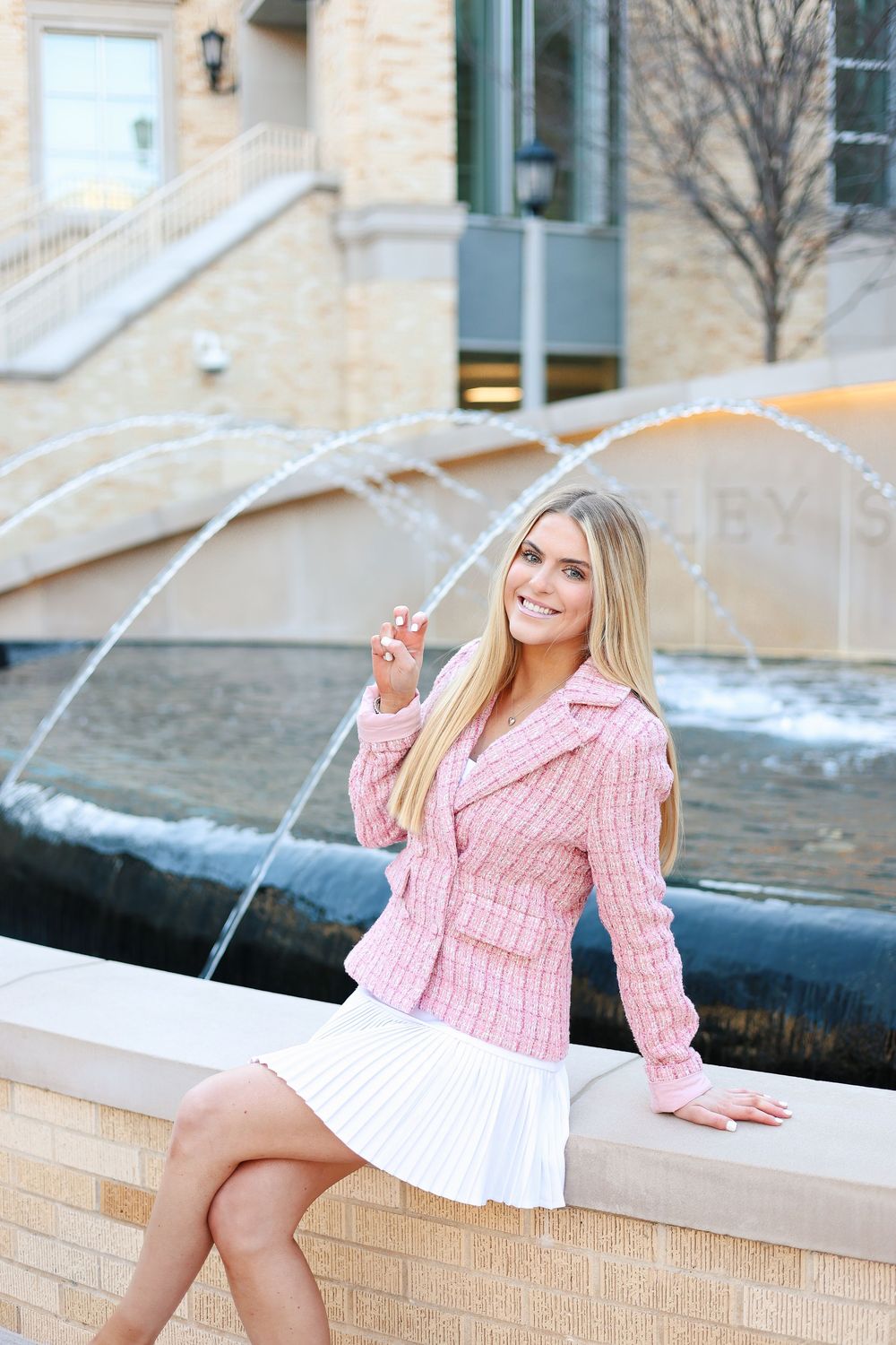 Fashion photoshoot by fountain featuring pink sweater and white skirt against stone wall backdrop.