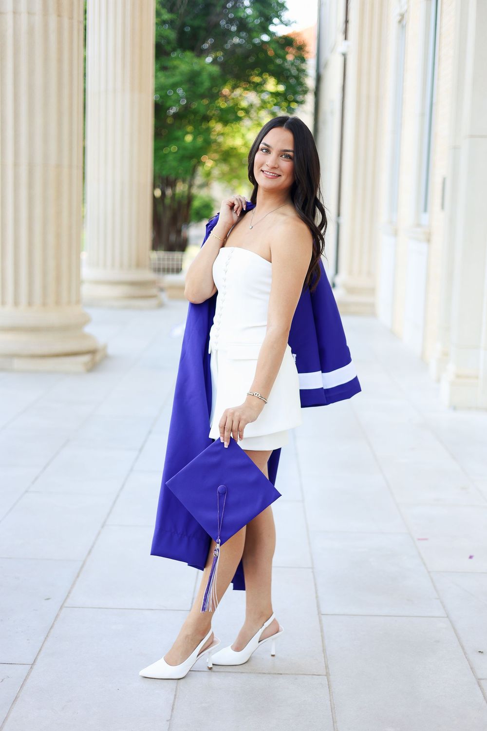 Graduate poses elegantly in white dress with blue graduation cap near classical columns.