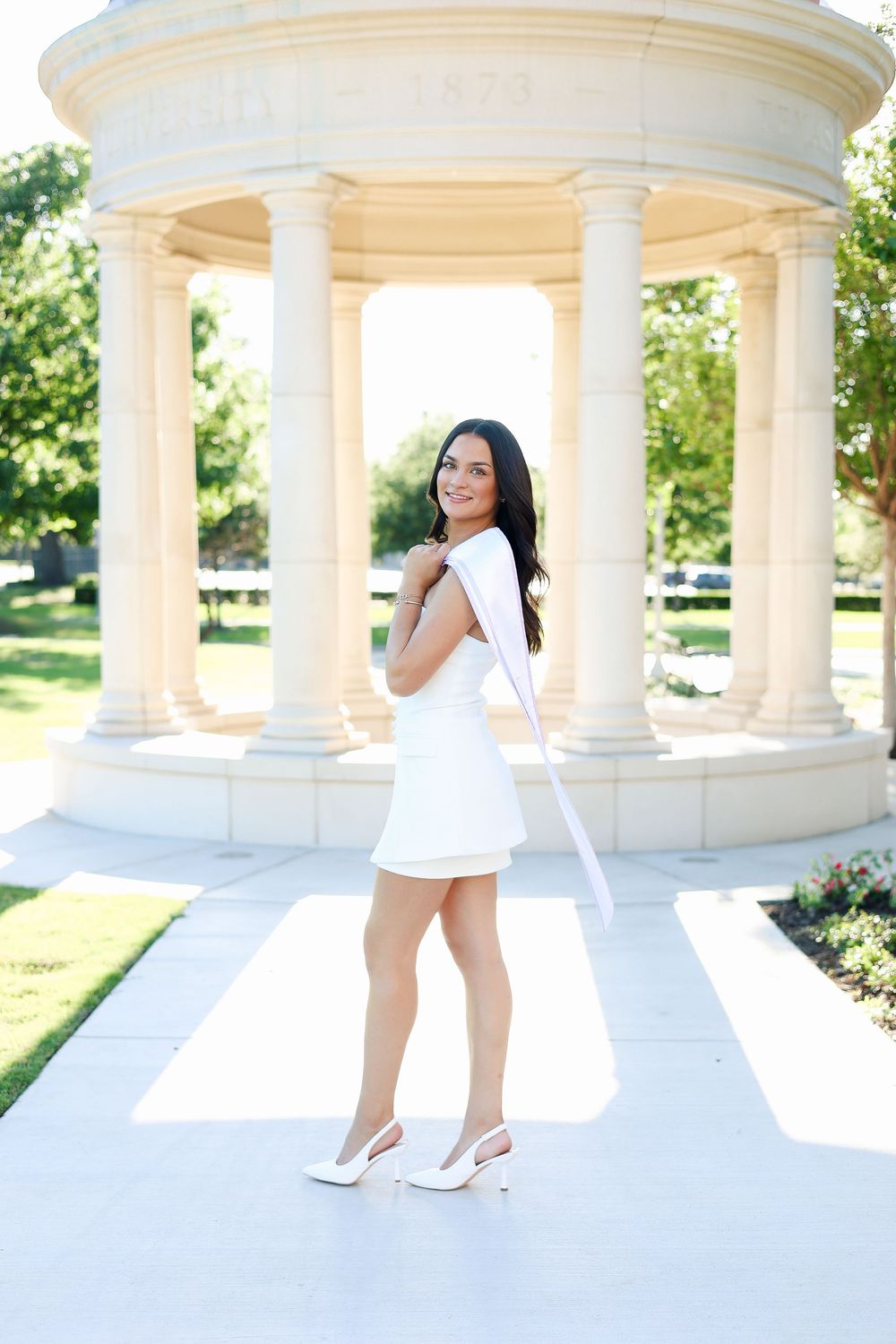 Graduate celebrates in white dress at ornate outdoor pavilion with classical white columns.