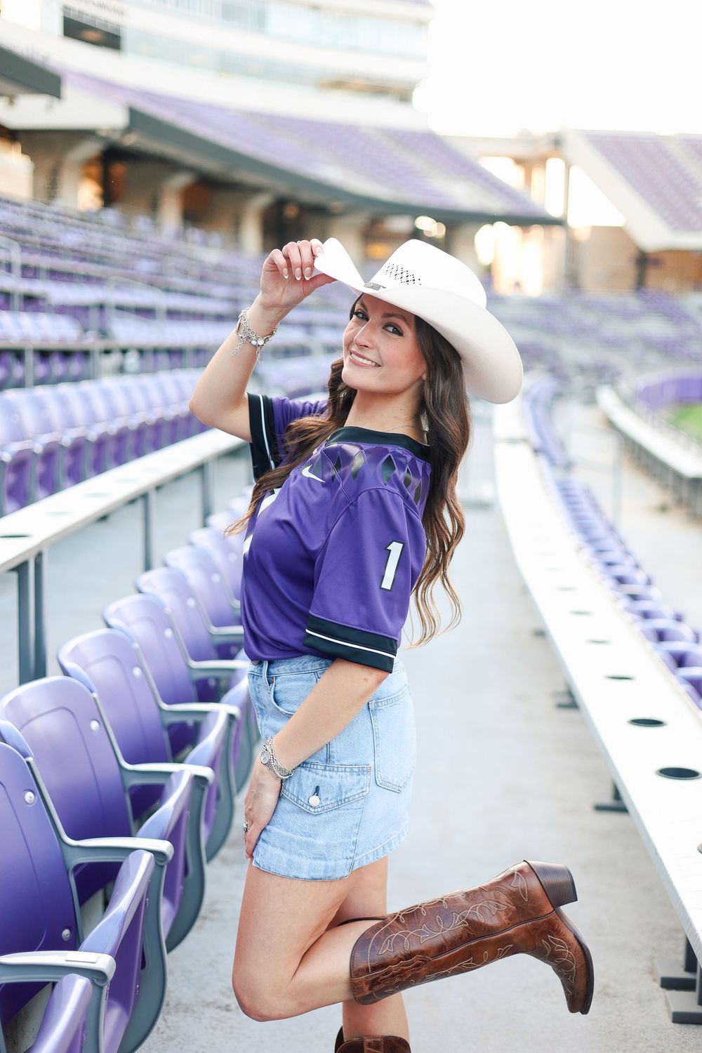 Graduate in purple jersey and cowboy boots celebrates in empty stadium seats.
