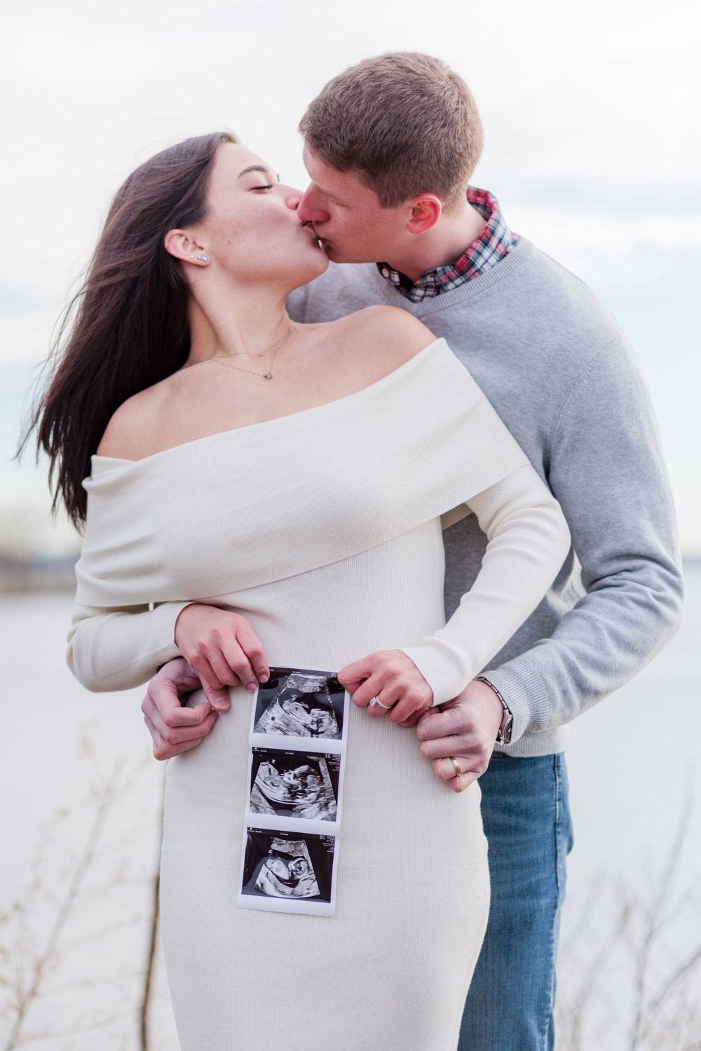 Husband and wife facing the camera, he is holding his pregnant wife. They are kissing with a sonogram photo held over the woman's belly.