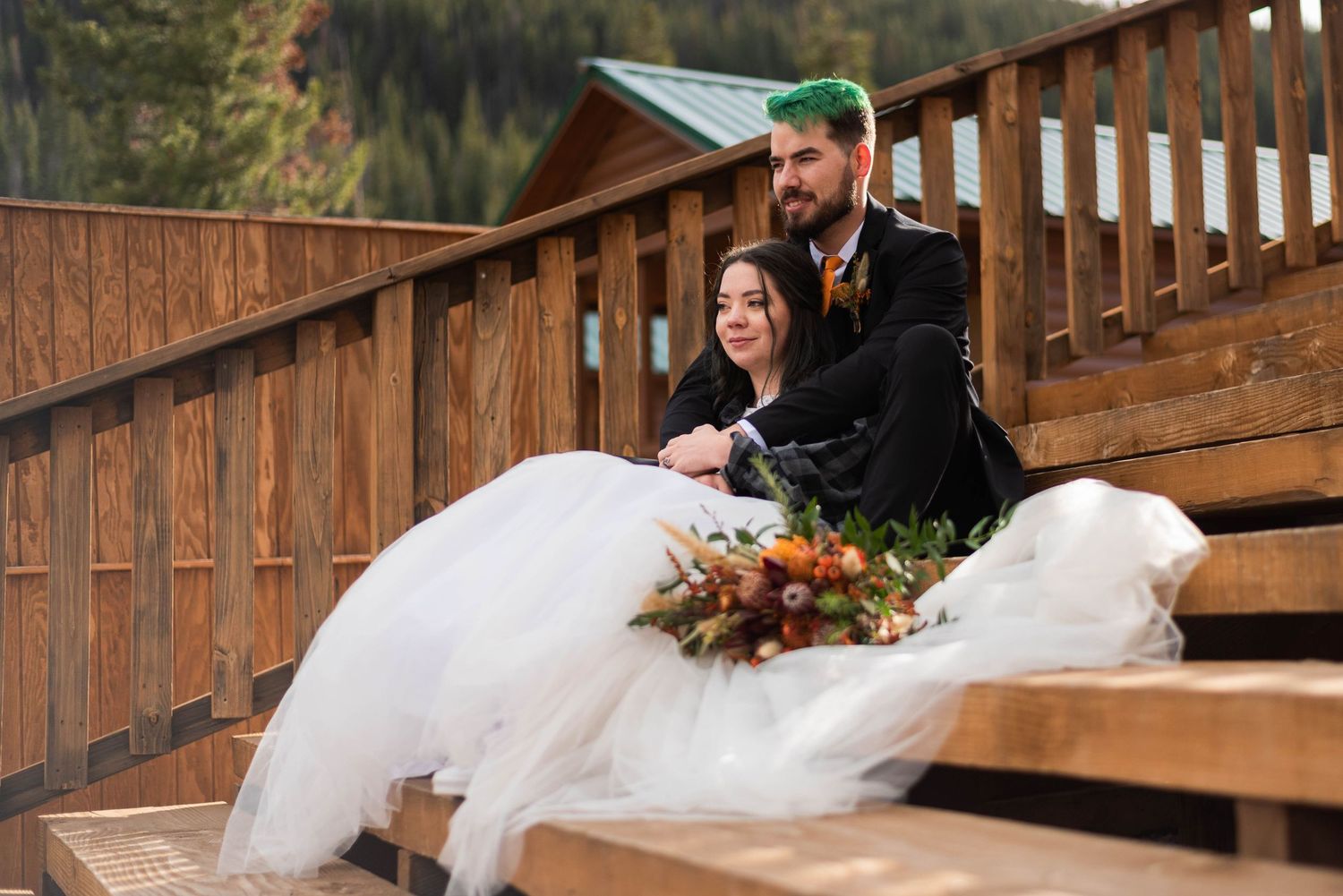 A couple sits together on wooden stairs outdoors, with a flowing white wedding dress and autumn bouquet.