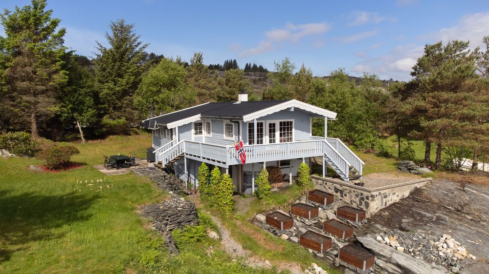Gray wooden cabin with deck and stairs surrounded by lush green trees and rocky shoreline on a sunny day.