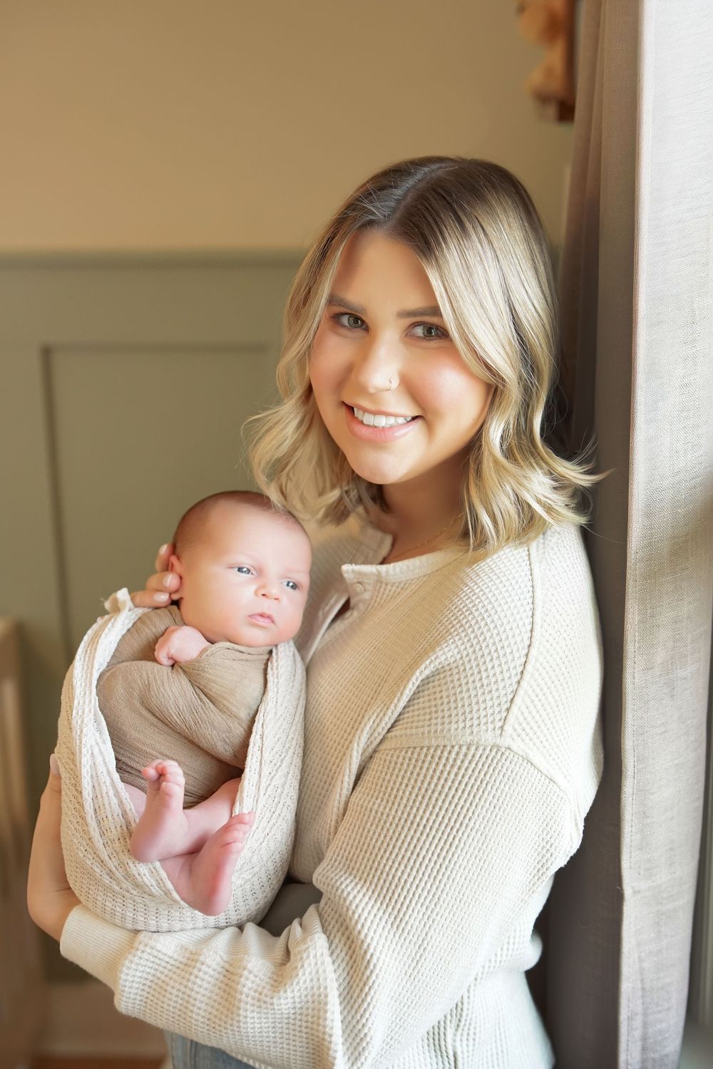 Smiling blonde mother in a cream knit sweater holds her newborn baby wrapped in a soft beige swaddle.