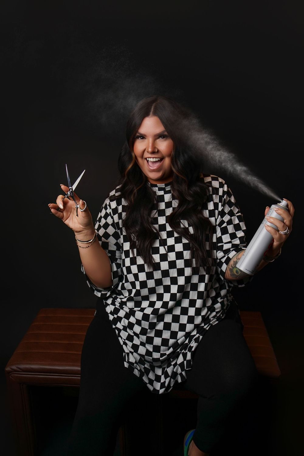 Smiling hairstylist in a black and white checkered cape holding scissors and spraying hairspray against a dark studio backdrop.